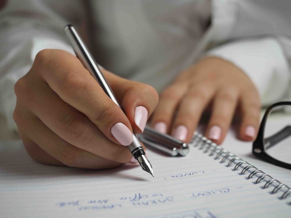 Woman in White Long Sleeved Shirt Holding a Pen Writing on a Paper | luxylounge